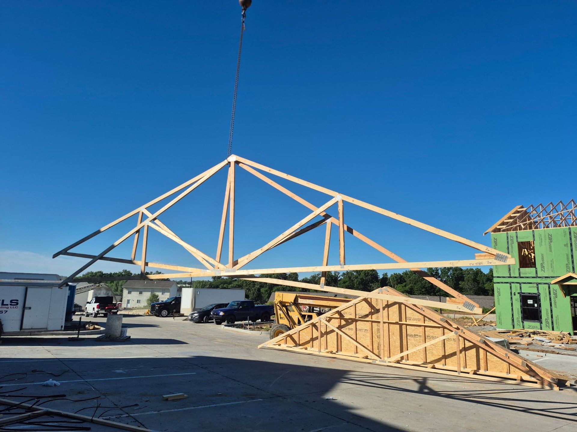 Construction site with large wooden roof truss being lifted by crane under a clear blue sky