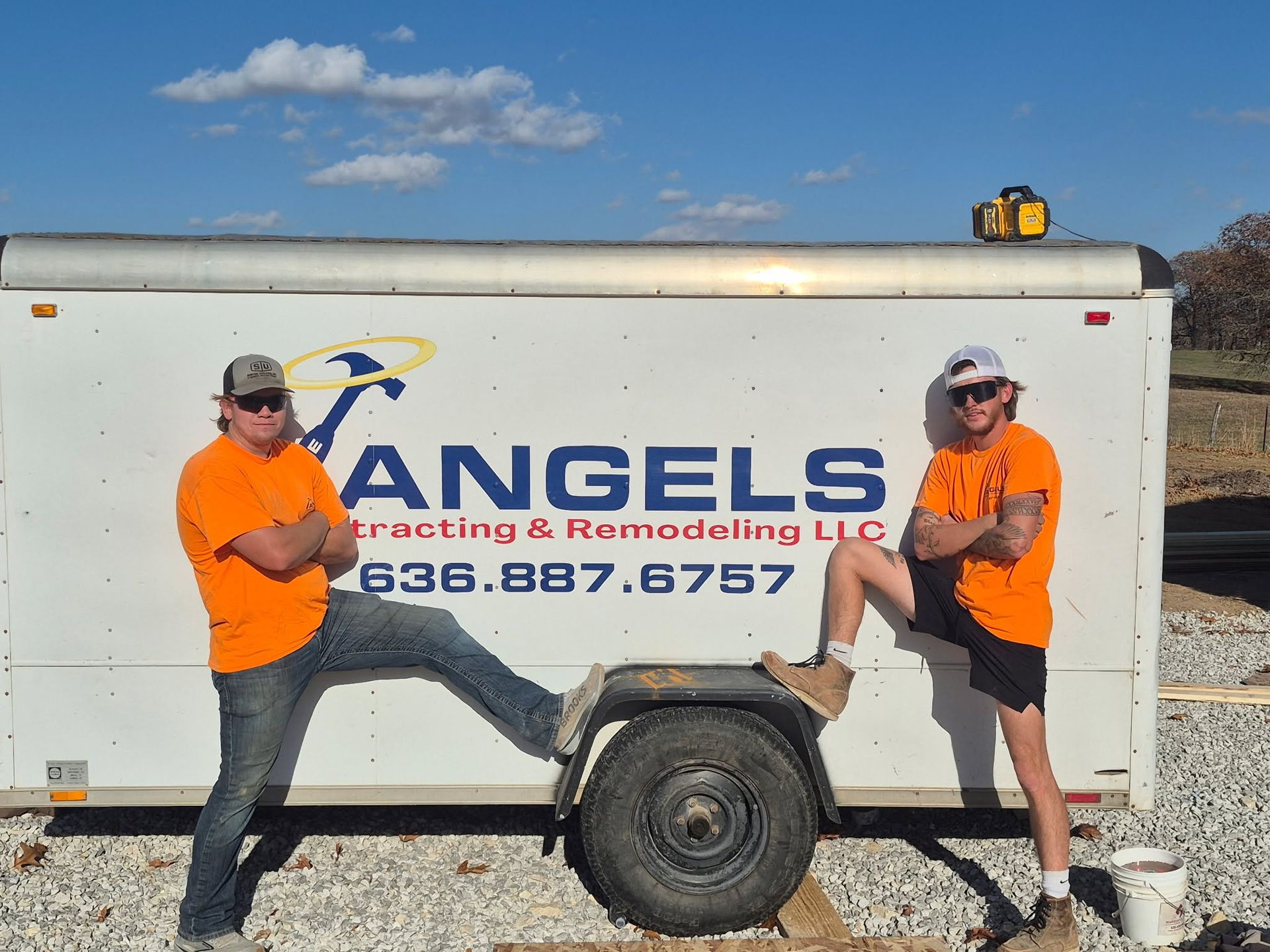 Two people in orange shirts posing beside an ANGELS Roofing & Remodeling LLC trailer on a sunny roadside.