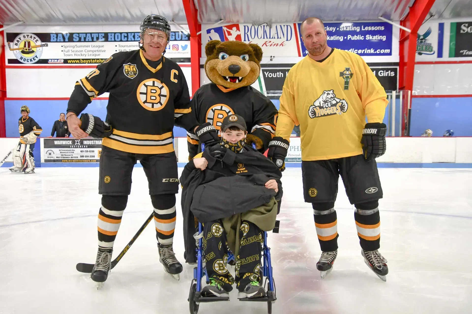Two hockey players, a mascot, and a child in a wheelchair on an ice rink, all wearing Bruins jerseys.