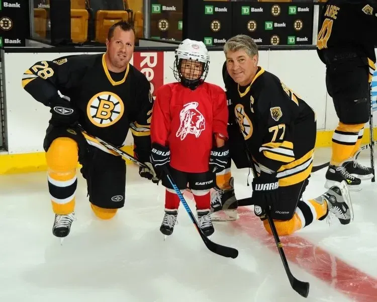 Two men in Bruins jerseys kneel with a child in a red jersey on an ice rink. They hold hockey sticks.