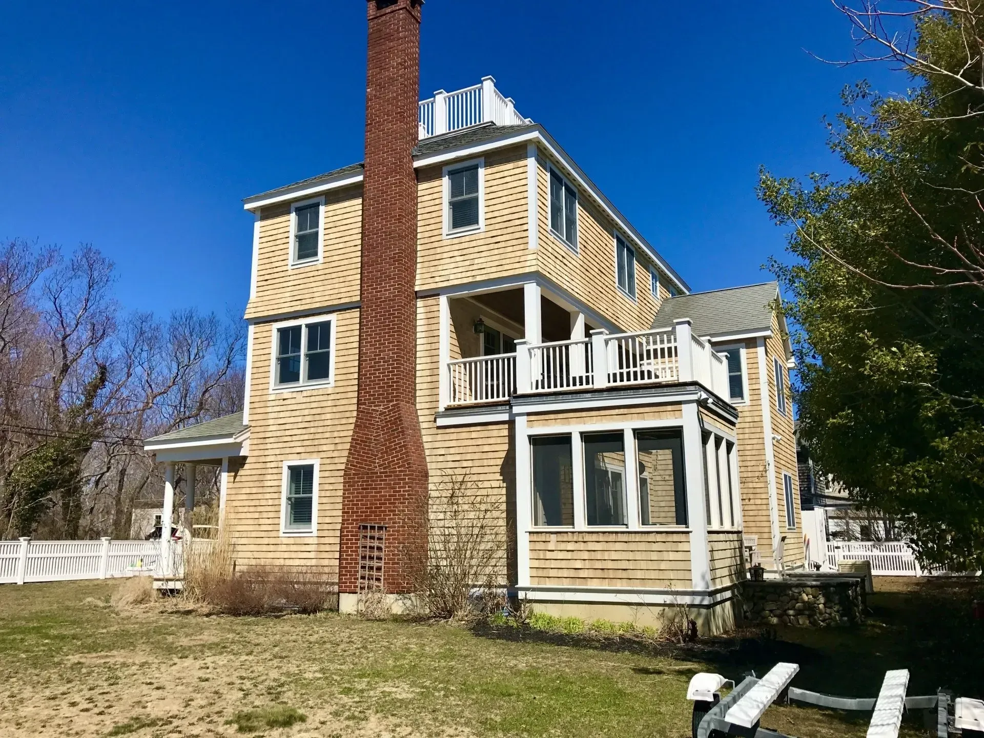 Three-story tan shingled house with a large brick chimney and white trim, sunny day.