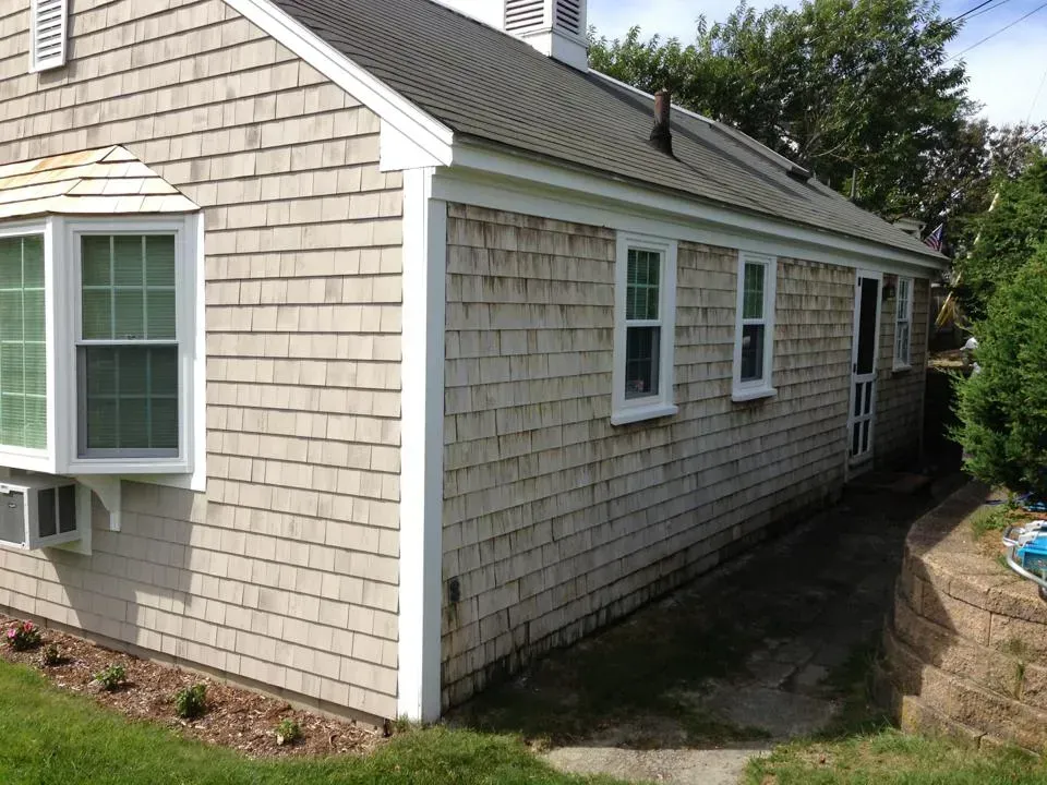 Shingled house with white trim, bay window, small windows, and dark roof on a sunny day.