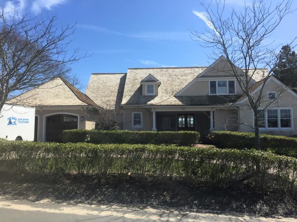 Beige house with wood shake roof, dormers, garage, and hedge row in front.