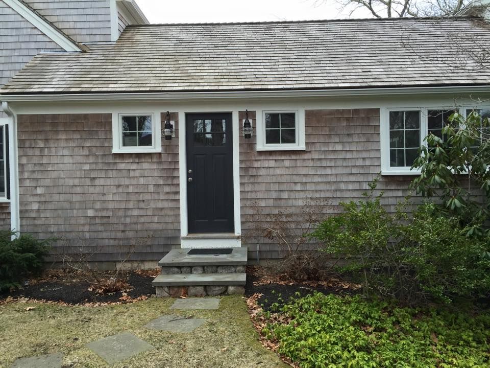 Gray shingled house with dark door and white-framed windows. Steps lead to the entrance, with some foliage.