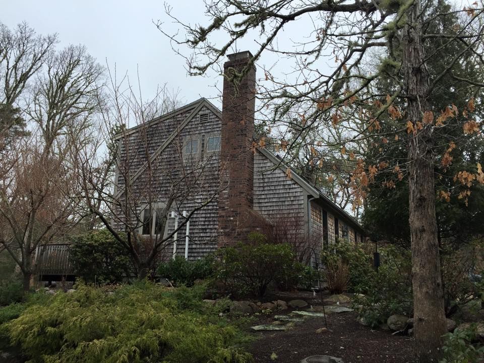 A weathered wooden house with a tall brick chimney, surrounded by trees and bushes.