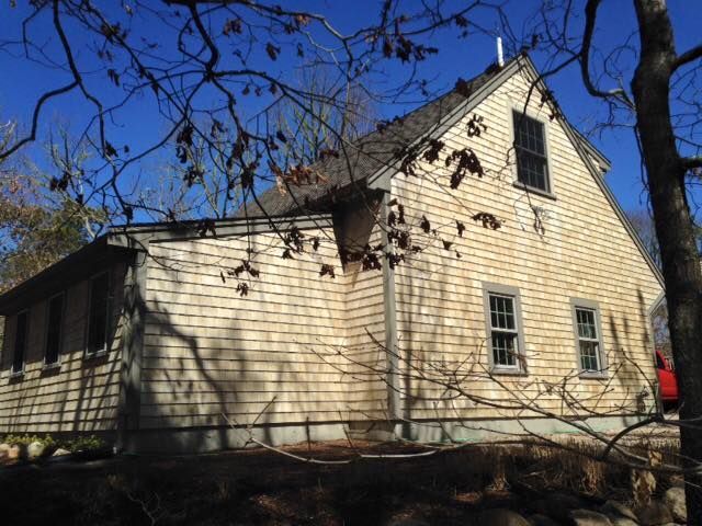 Beige shingled house with dark windows under a blue sky, partially obscured by tree branches.