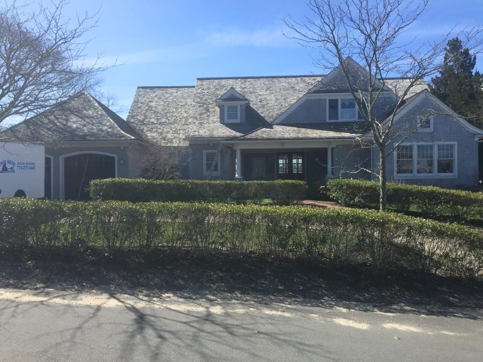 Gray house with shingle roof, dormer windows, and trimmed hedges in front.