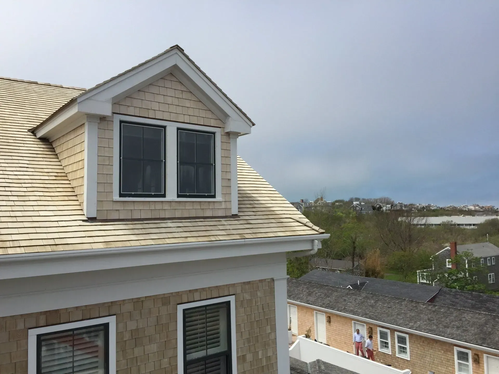 Dormer window on a light-colored shingle roof with a view of a town under a cloudy sky.