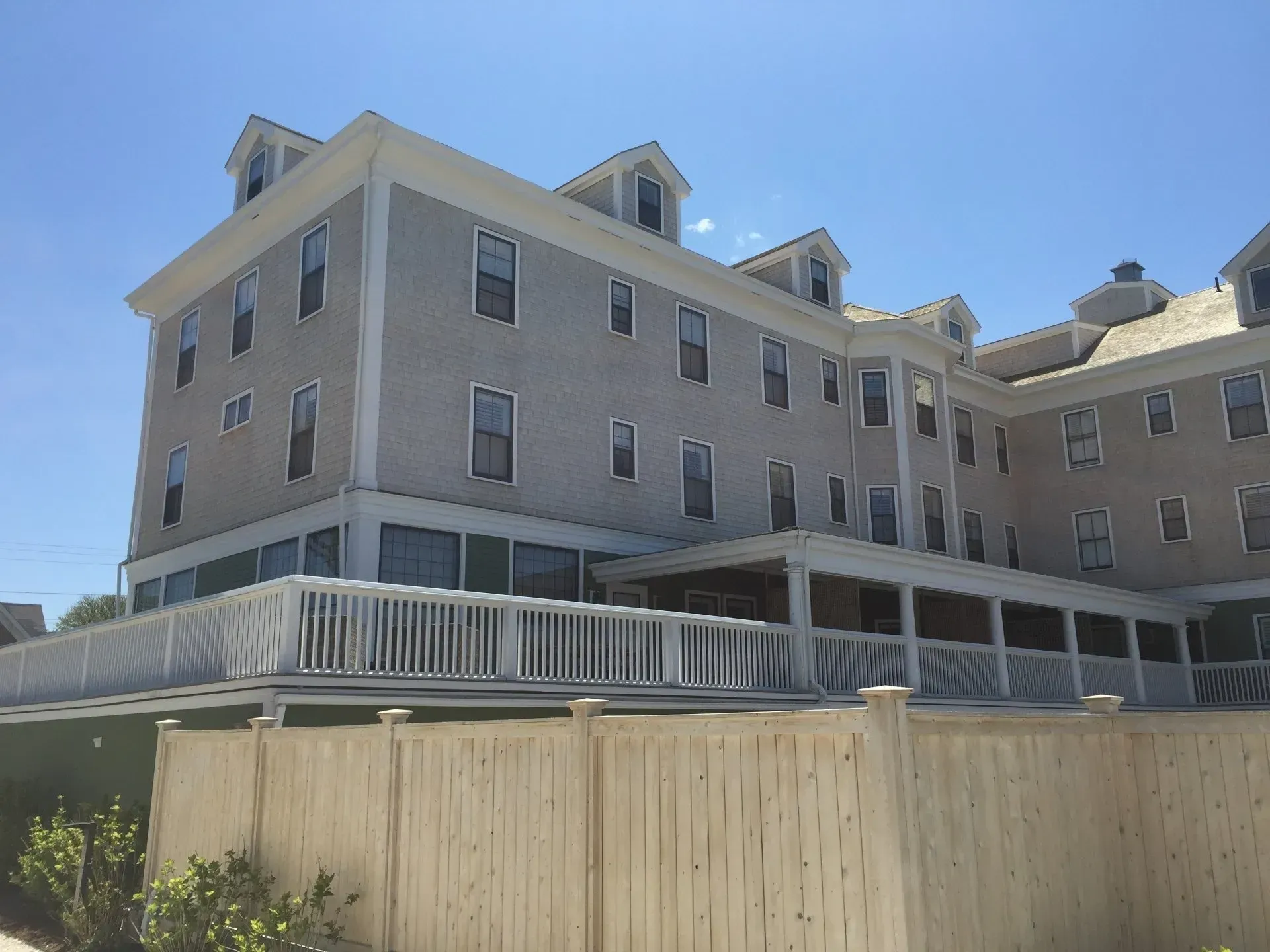 Three-story light gray building with dormers, a porch, and a wooden fence on a sunny day.