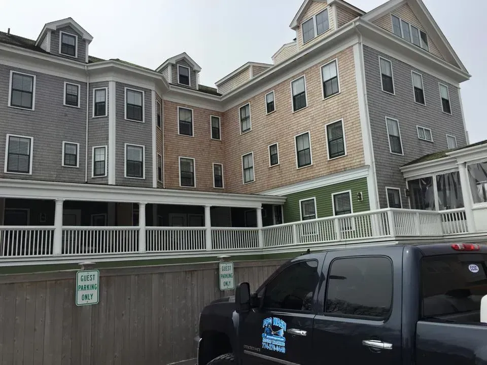 A multi-story building with gray, tan, and green siding, a wraparound porch, and a parked truck in front.