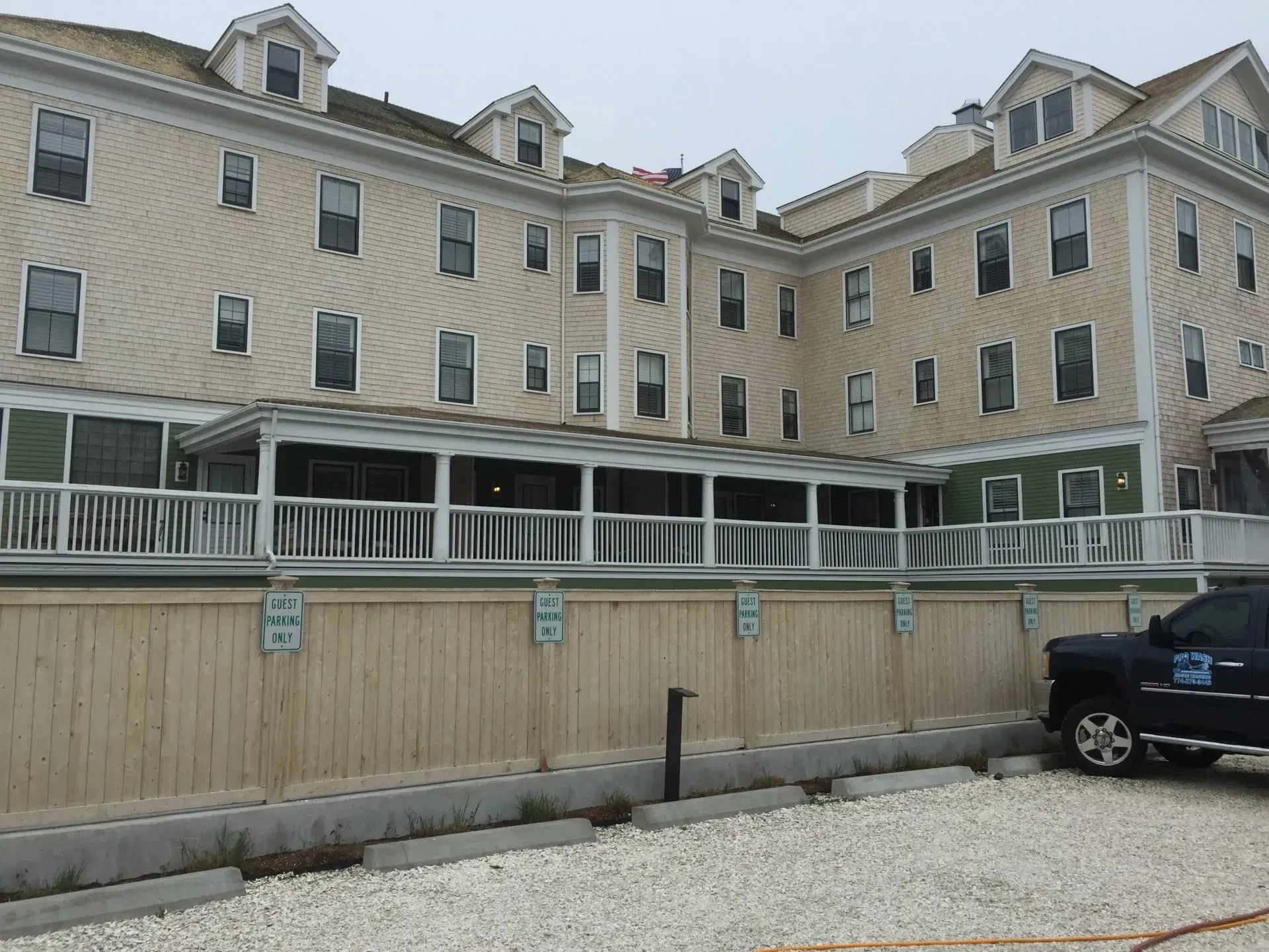 Tan multi-story building with a porch, surrounded by a wooden fence and parking lot, overcast day.