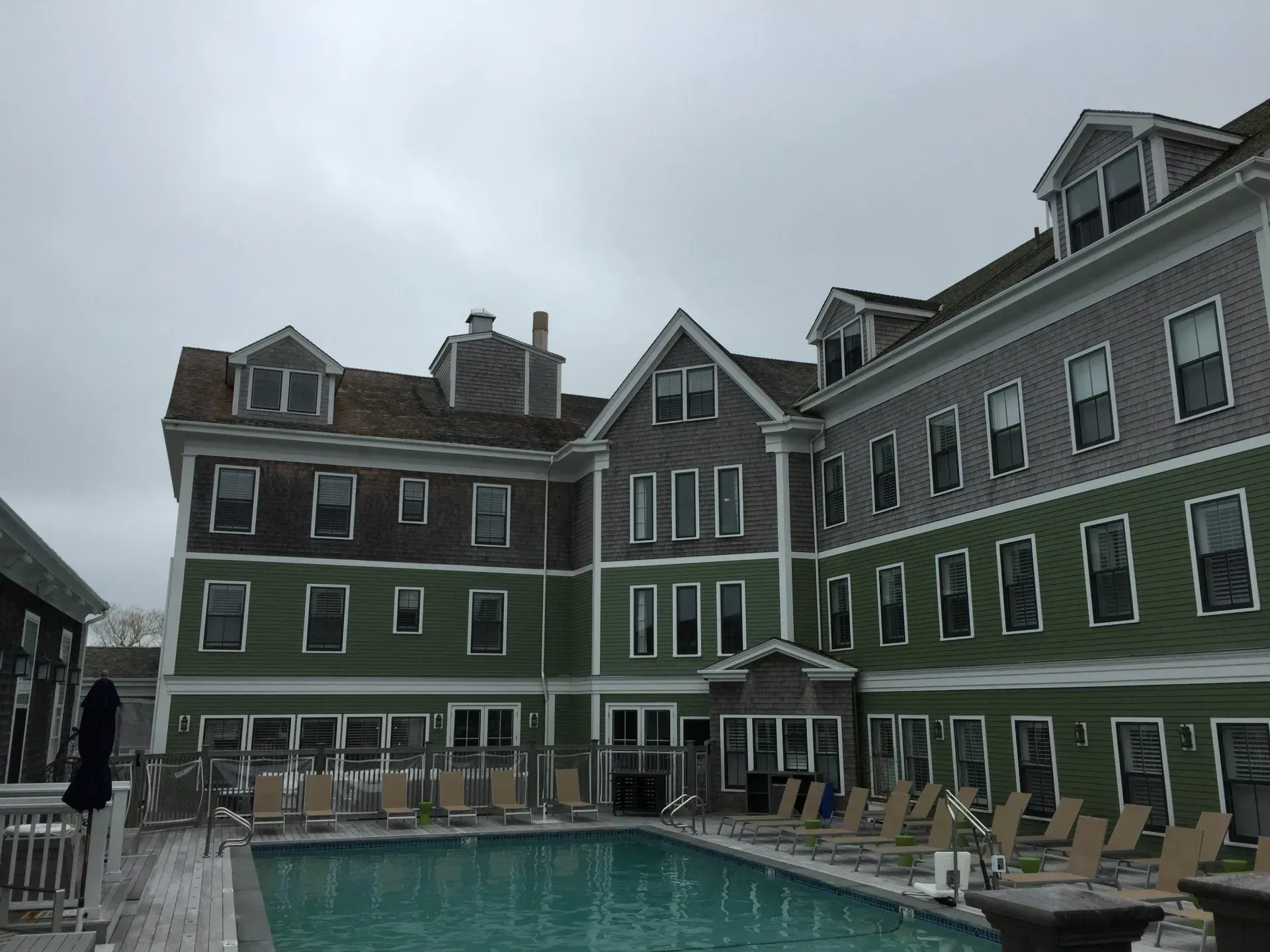 Hotel exterior with a swimming pool on an overcast day. Buildings are green and gray with multiple windows.