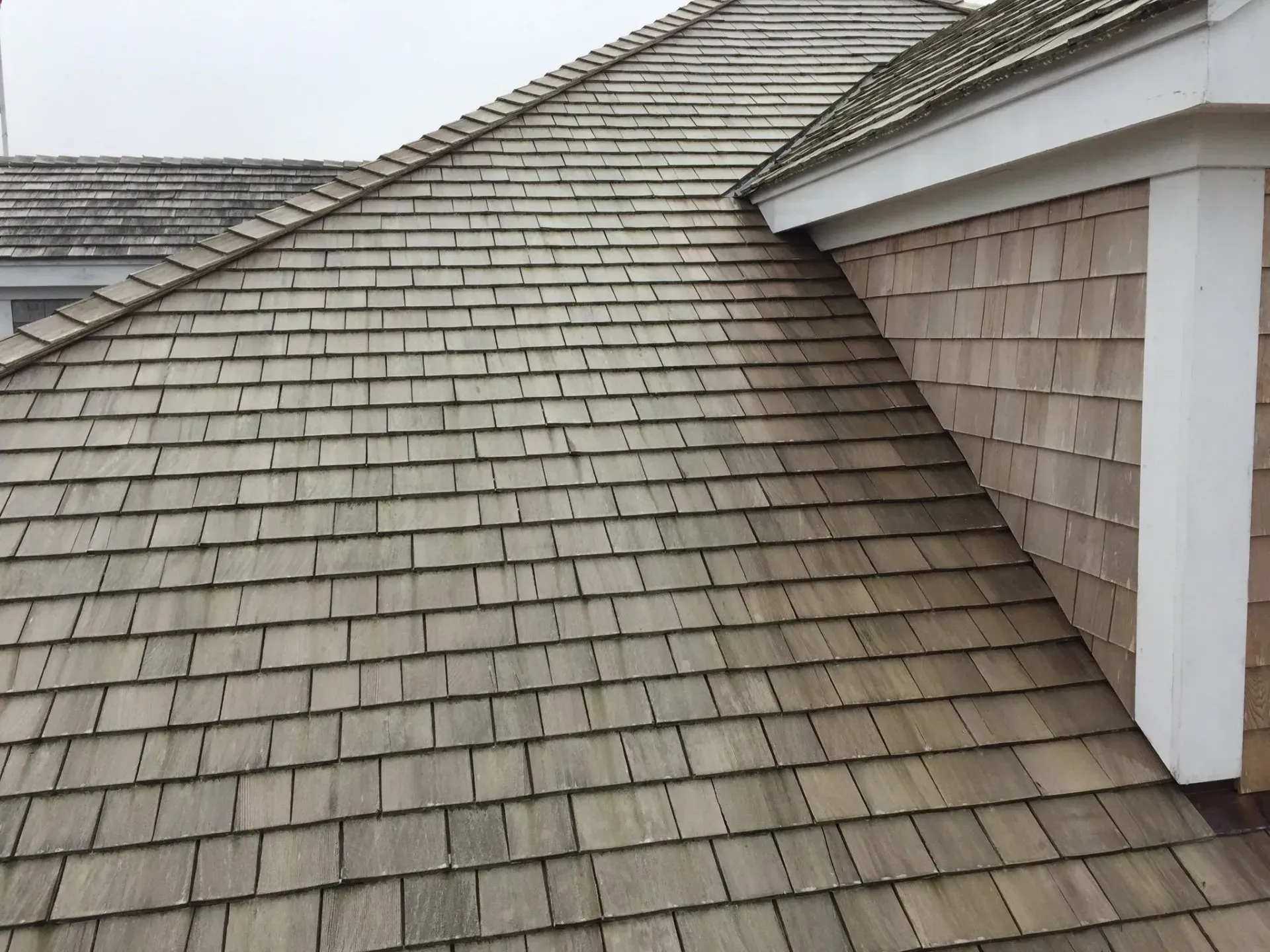 Wooden shingle roof with faded grey-brown color. Includes part of a white-trimmed corner and wooden siding.