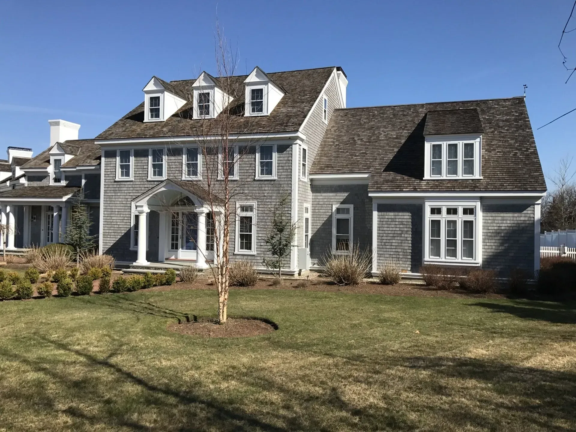 Two-story gray house with multiple dormers, white trim, and a small tree in front.