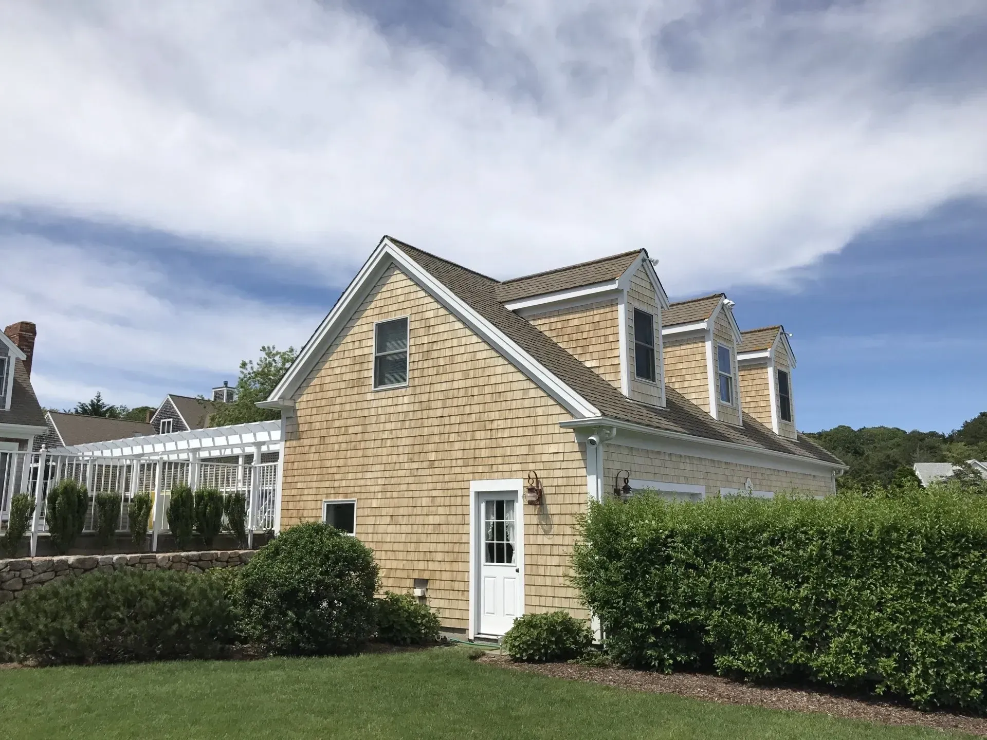 Tan shingled building with white trim, three dormers, and a white door, surrounded by green shrubs and lawn.