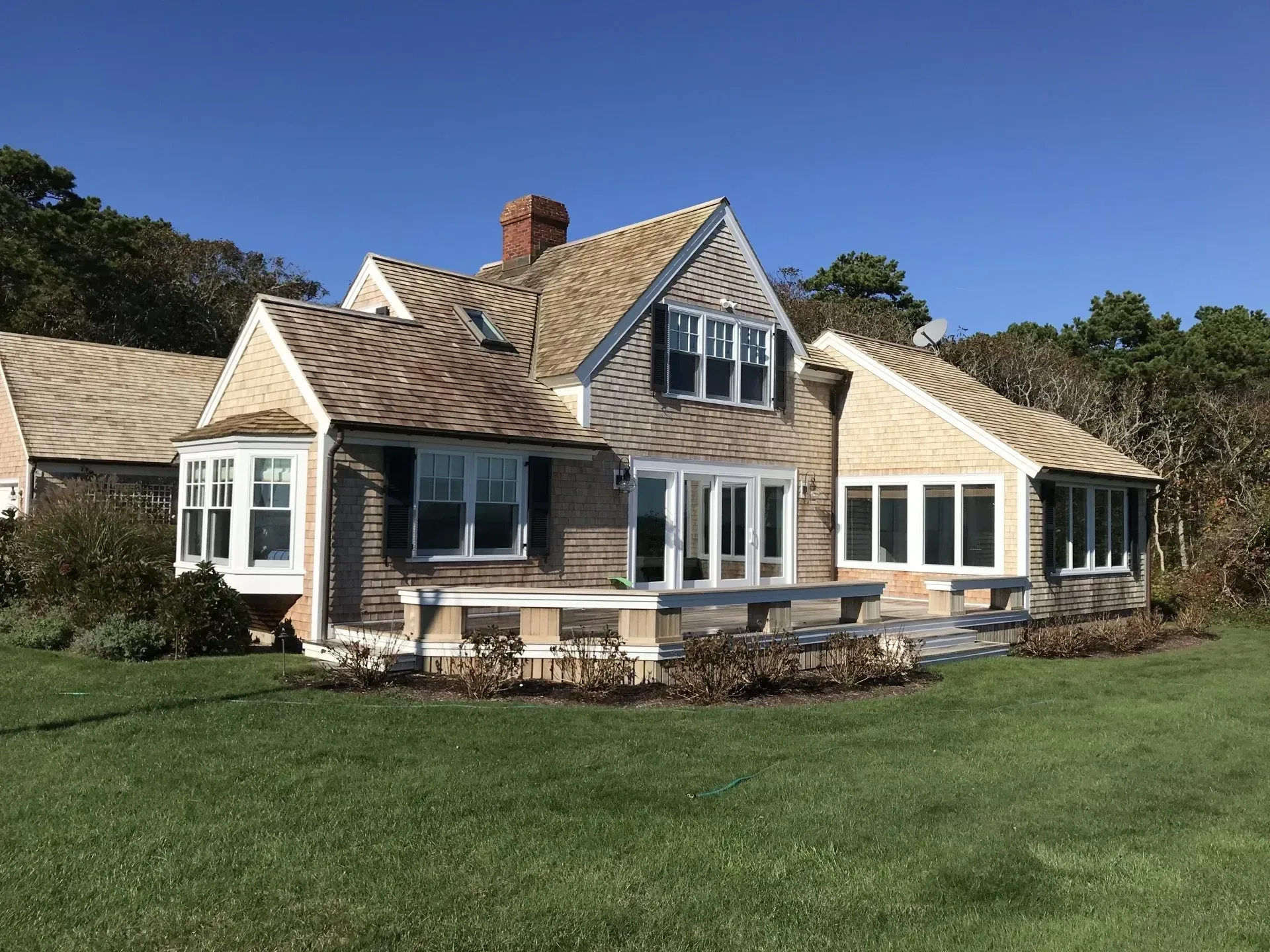 Cottage-style house with cedar shake roof, brown siding, and large windows on a green lawn against a clear blue sky.