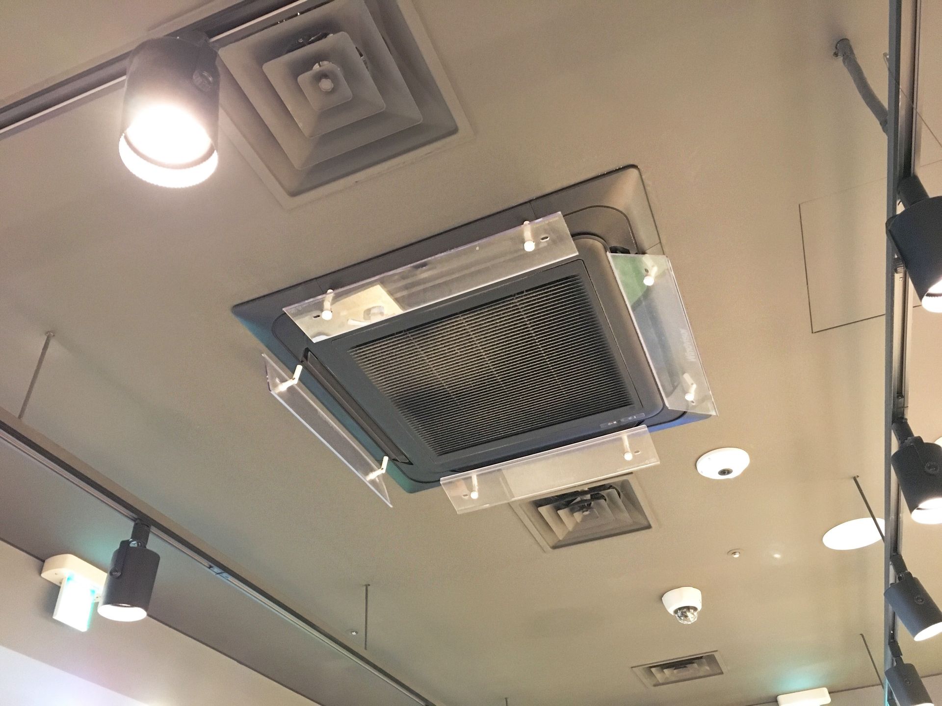 Ceiling-mounted air conditioner unit with protective plastic shields, recessed lighting, and a security camera.