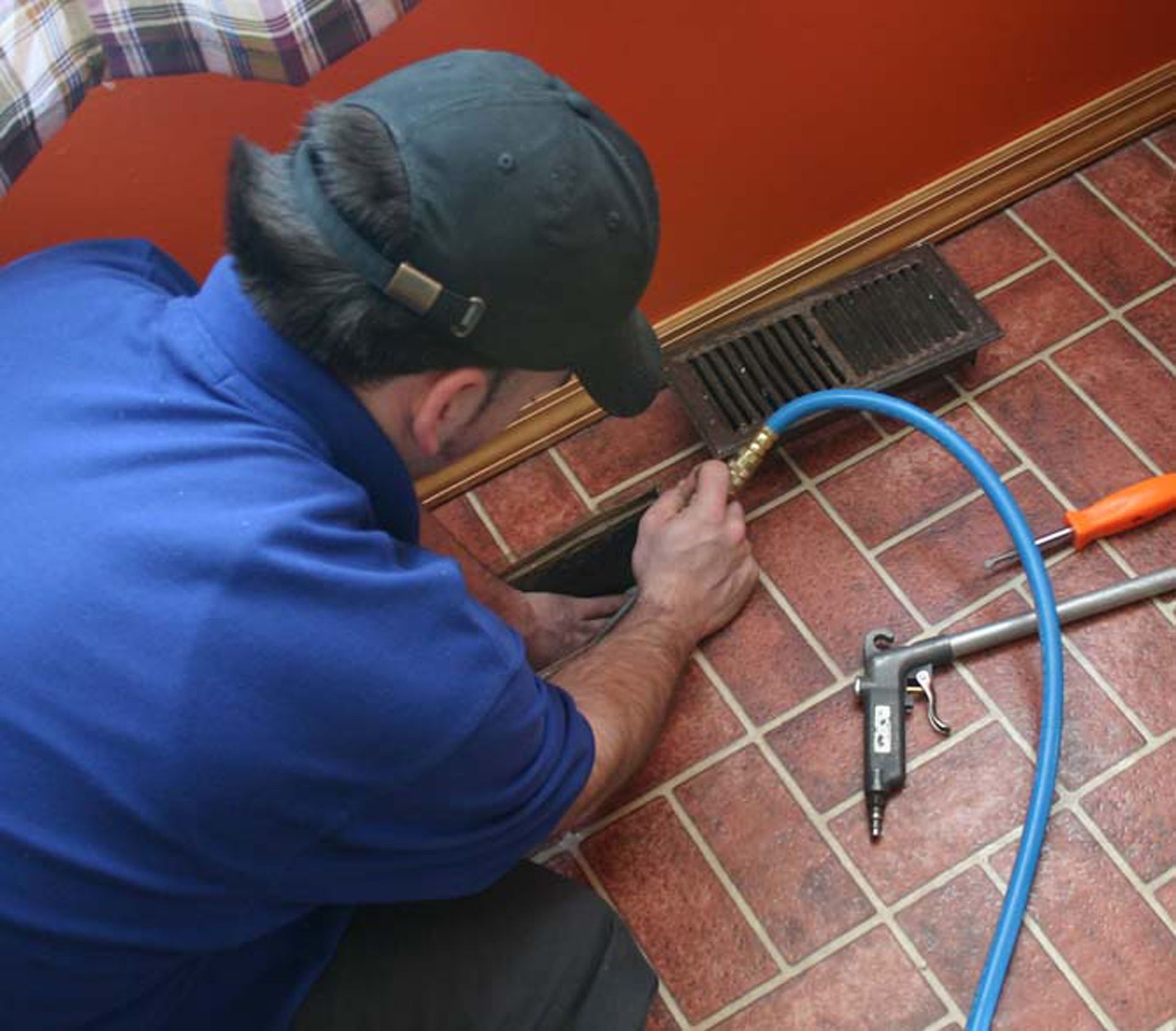 Man in blue shirt cleaning floor vent with air hose on a brick-like floor.