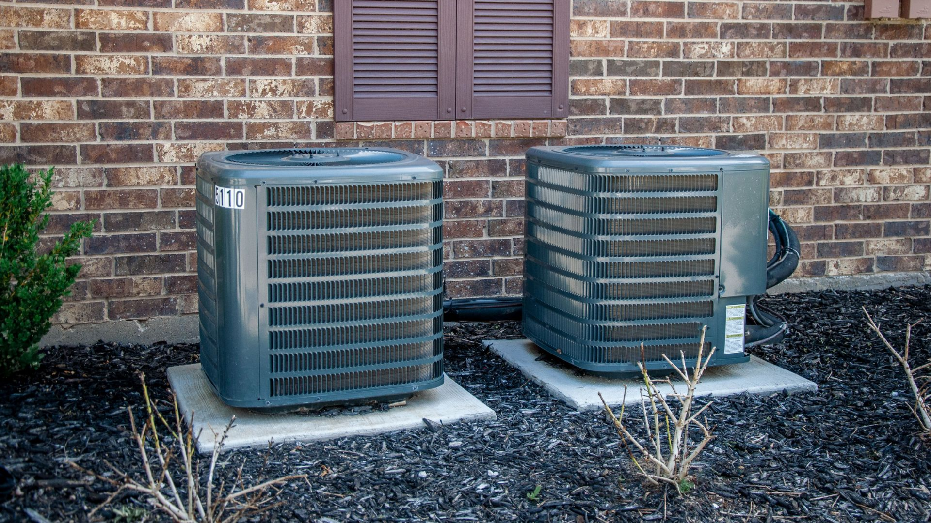 Two air conditioning units on concrete pads in front of a brick building with a brown shutter.