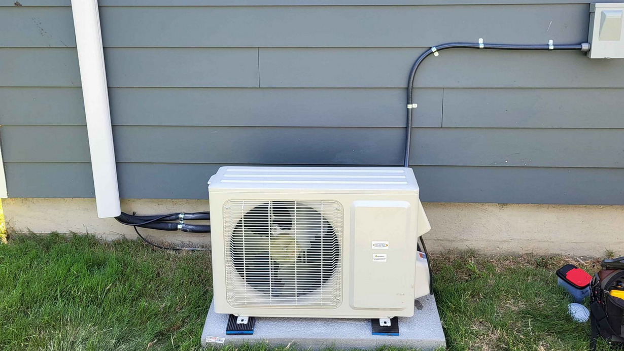 Outdoor air conditioning unit against a blue-gray house, on a concrete block, with black pipes and wiring.