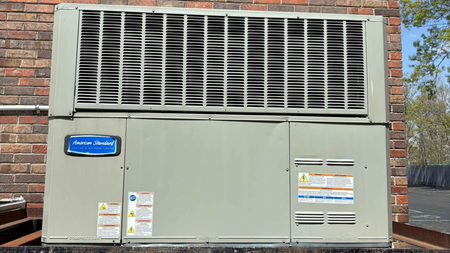 A gray American Standard air conditioning unit against a brick wall.