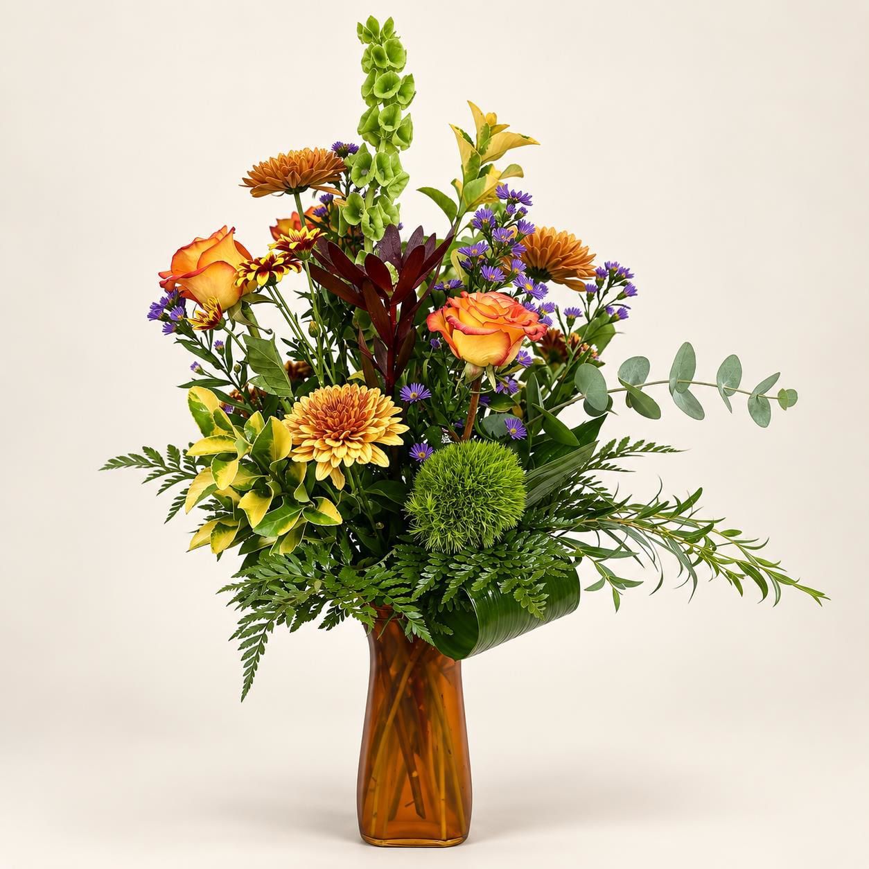 Colorful mixed bouquet of orange, yellow, green, and purple flowers in a glass vase on a light background