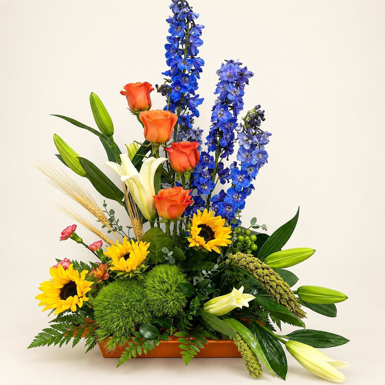 Colorful floral arrangement with blue delphiniums, orange roses, yellow sunflowers, and green foliage in a vase