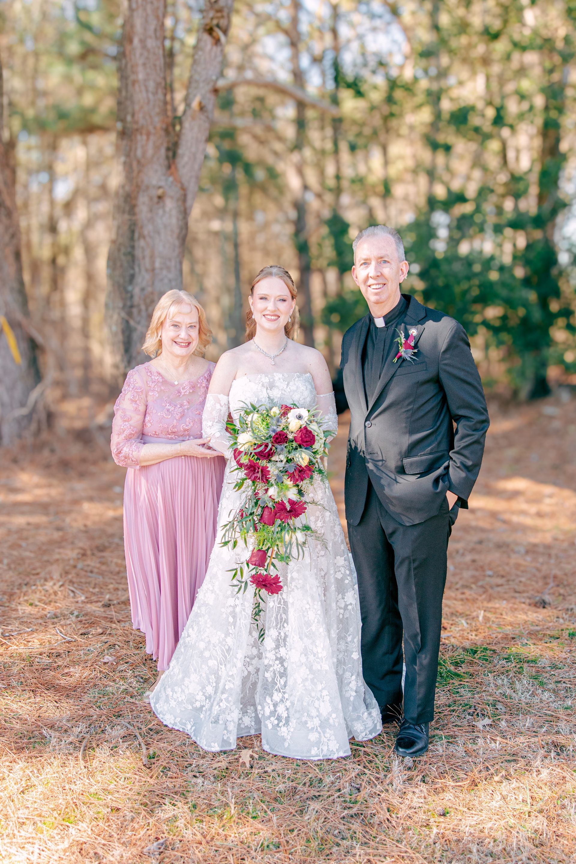 Three people posing outdoors at a wedding, with the center person holding a bouquet.