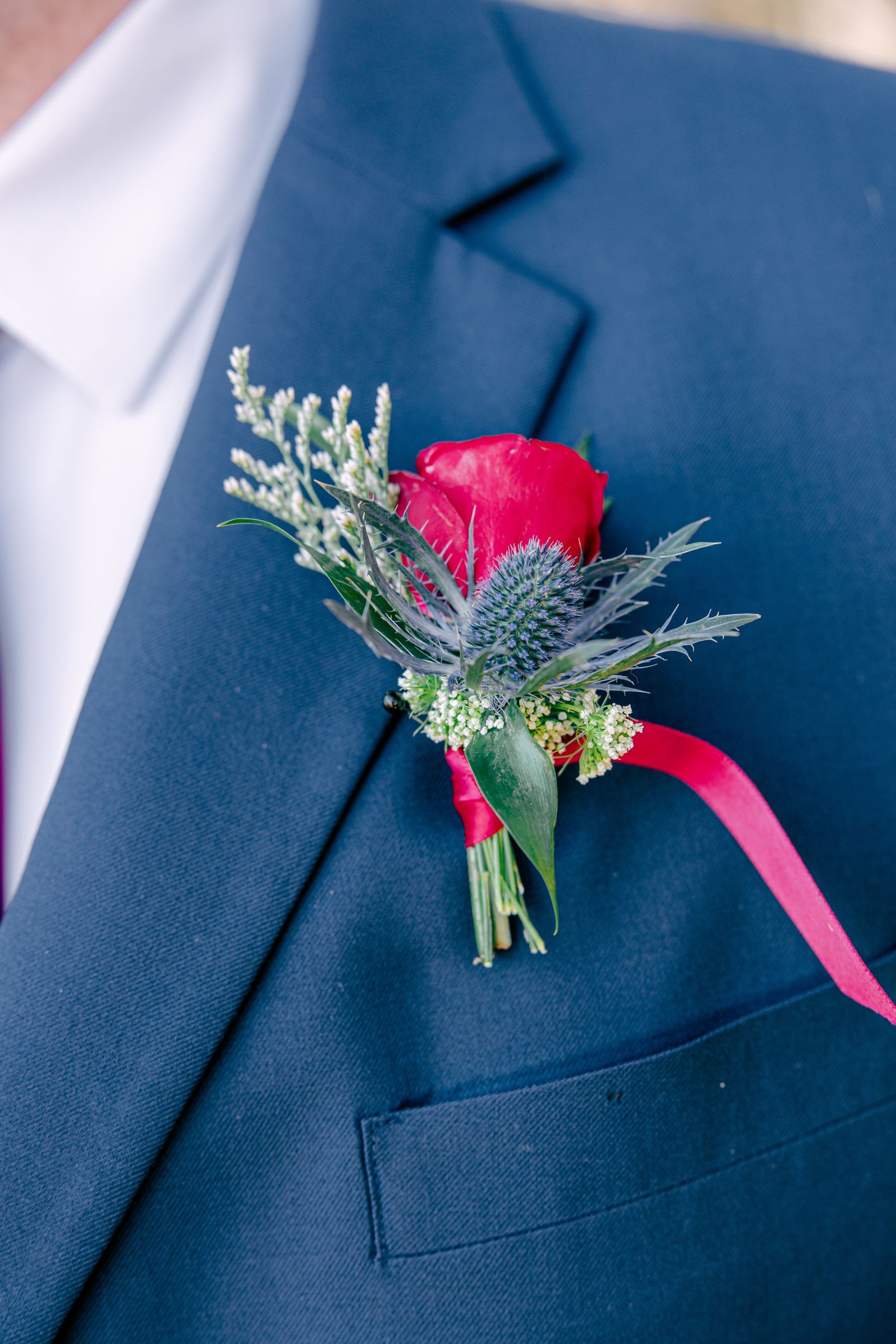 Red rose boutonniere with greenery and pink ribbon pinned to a navy suit jacket
