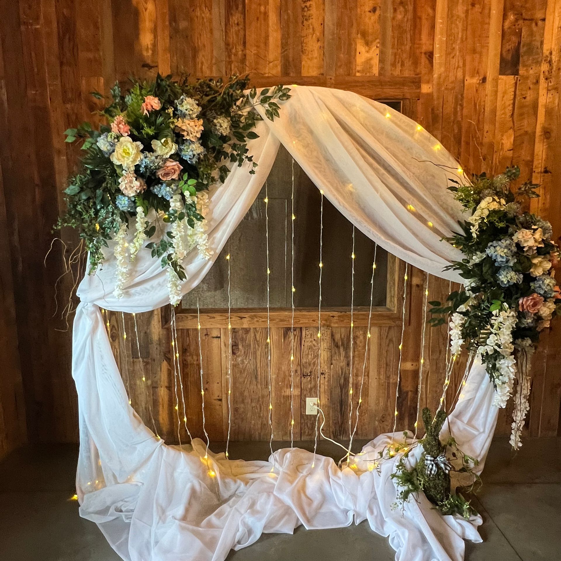 White draped wedding arch with floral arrangements against a rustic wood wall and fairy lights