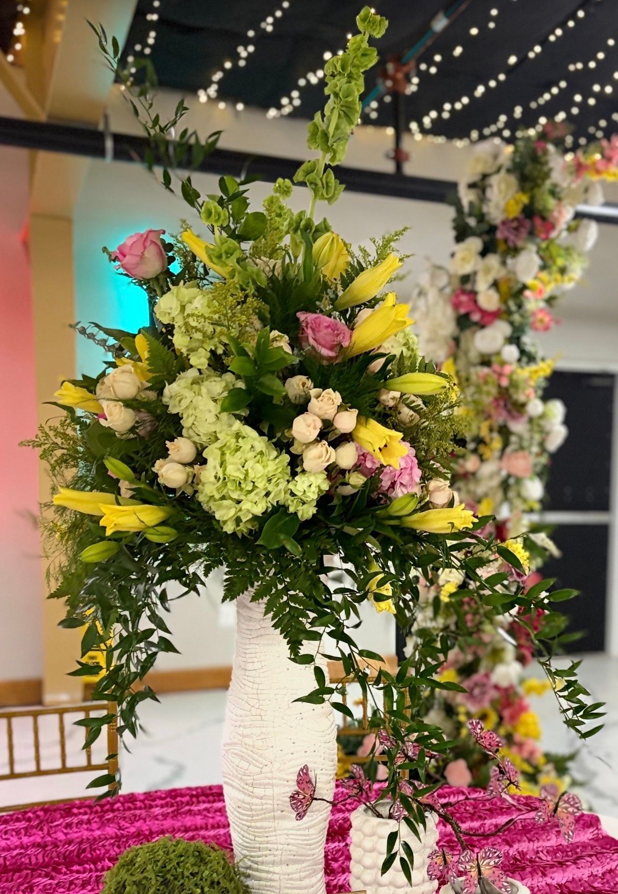 Tall floral arrangement with yellow, white, pink, and green flowers at an indoor event