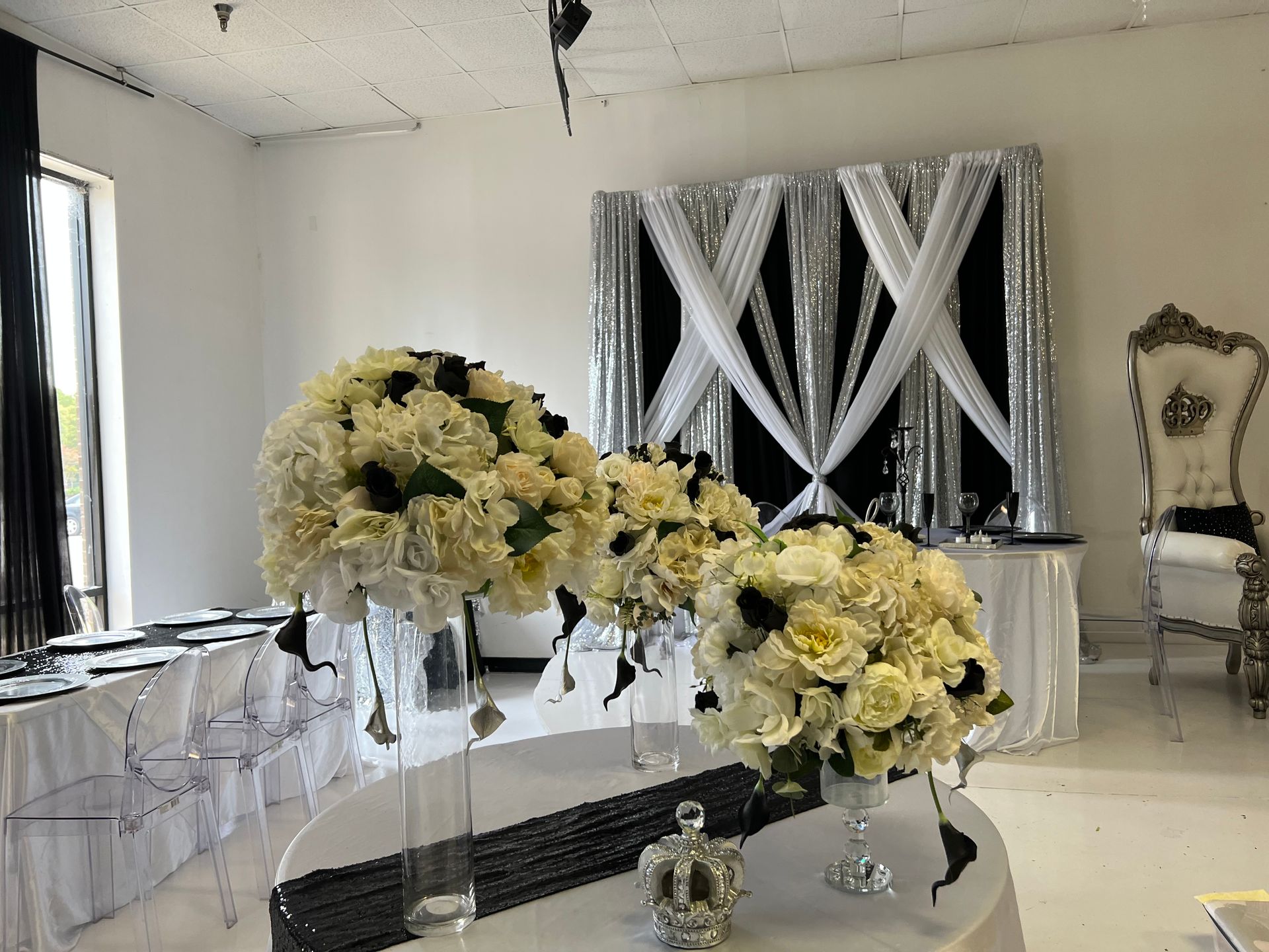 Elegant white wedding reception setup with floral centerpieces, draped backdrop, and clear chairs in a bright room