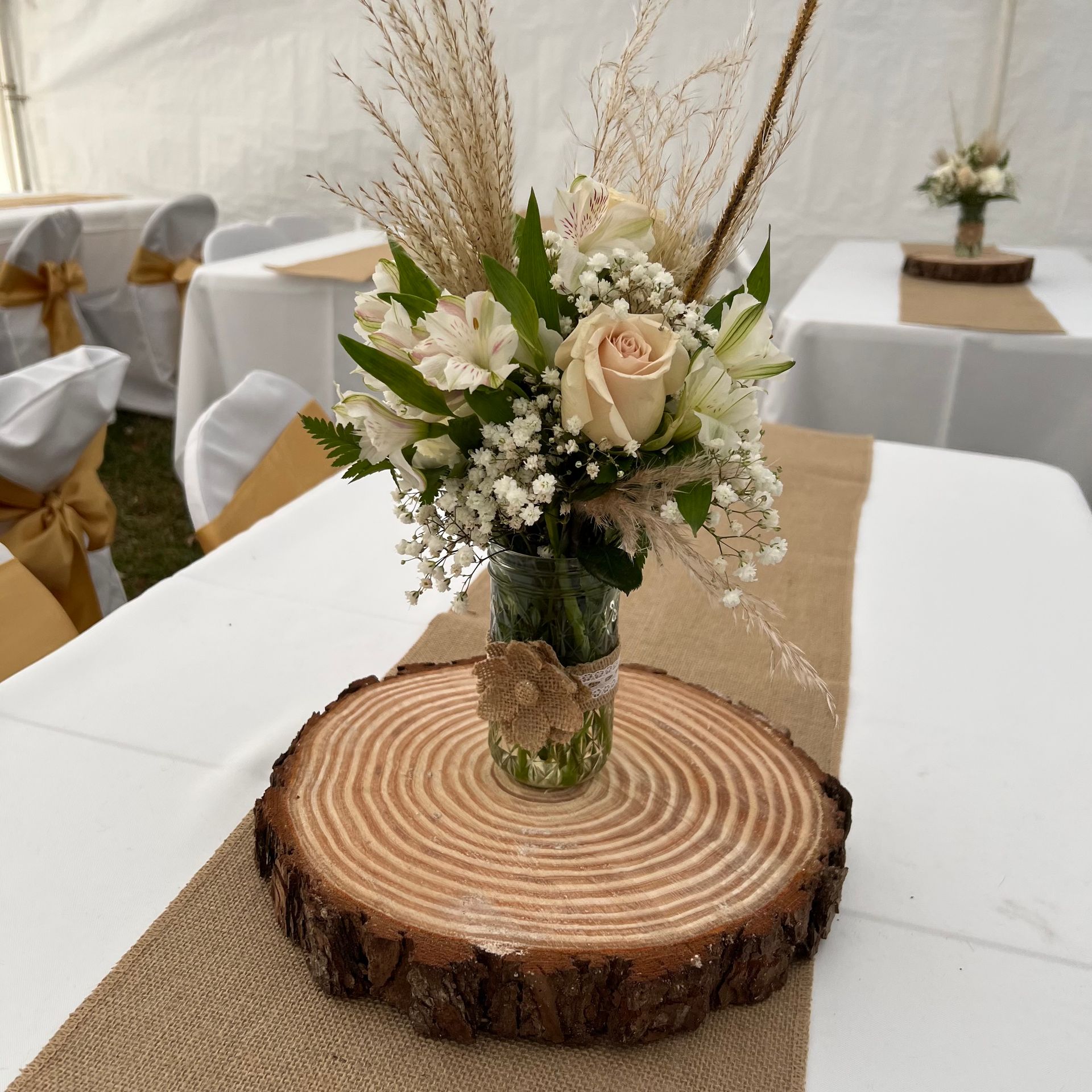 Rustic floral centerpiece in a glass vase on a wood-slice base, on a table in a decorated event venue.