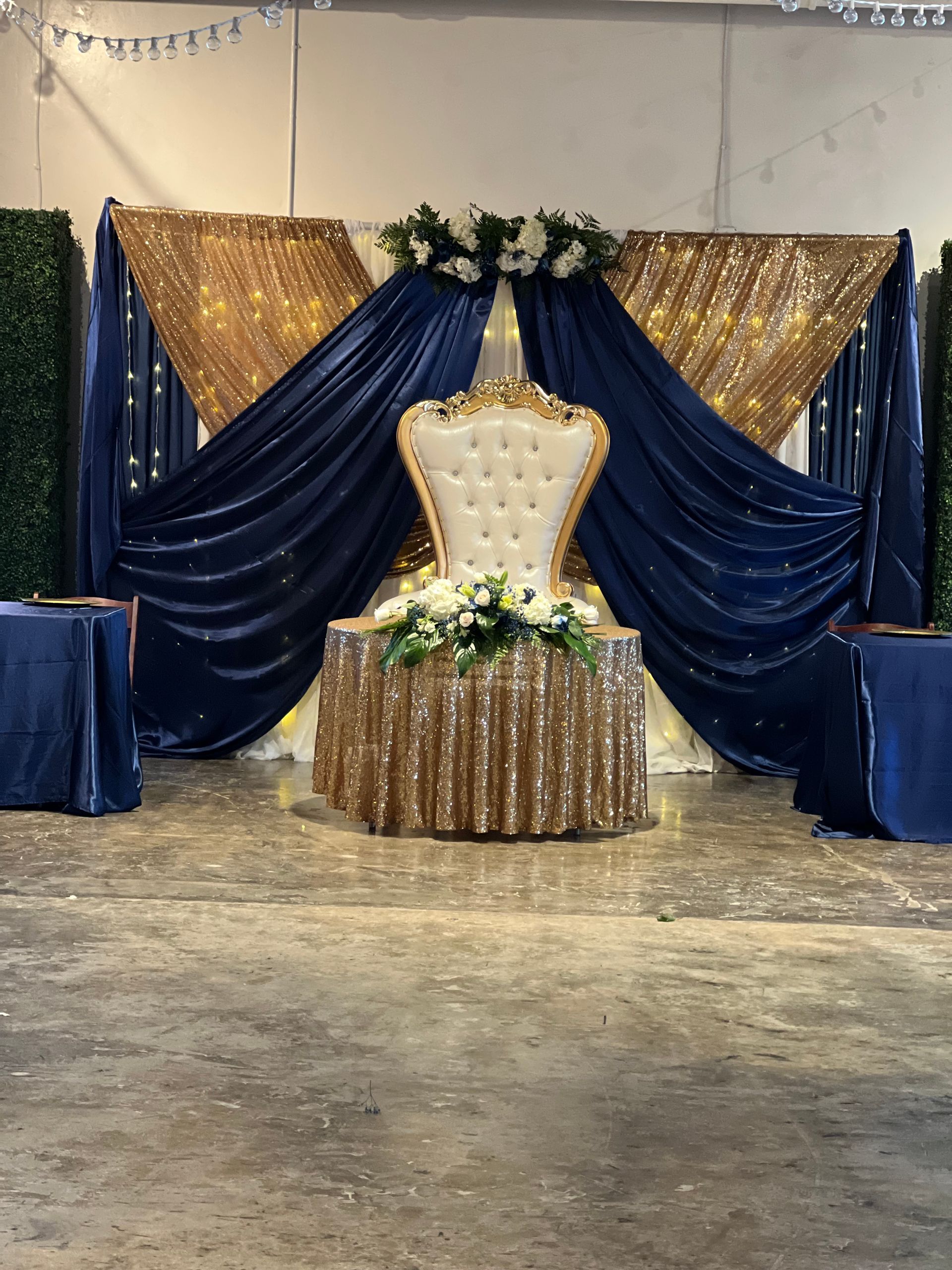 Gold throne chair on a decorated stage with navy blue and gold drapes, floral accents, and side tables