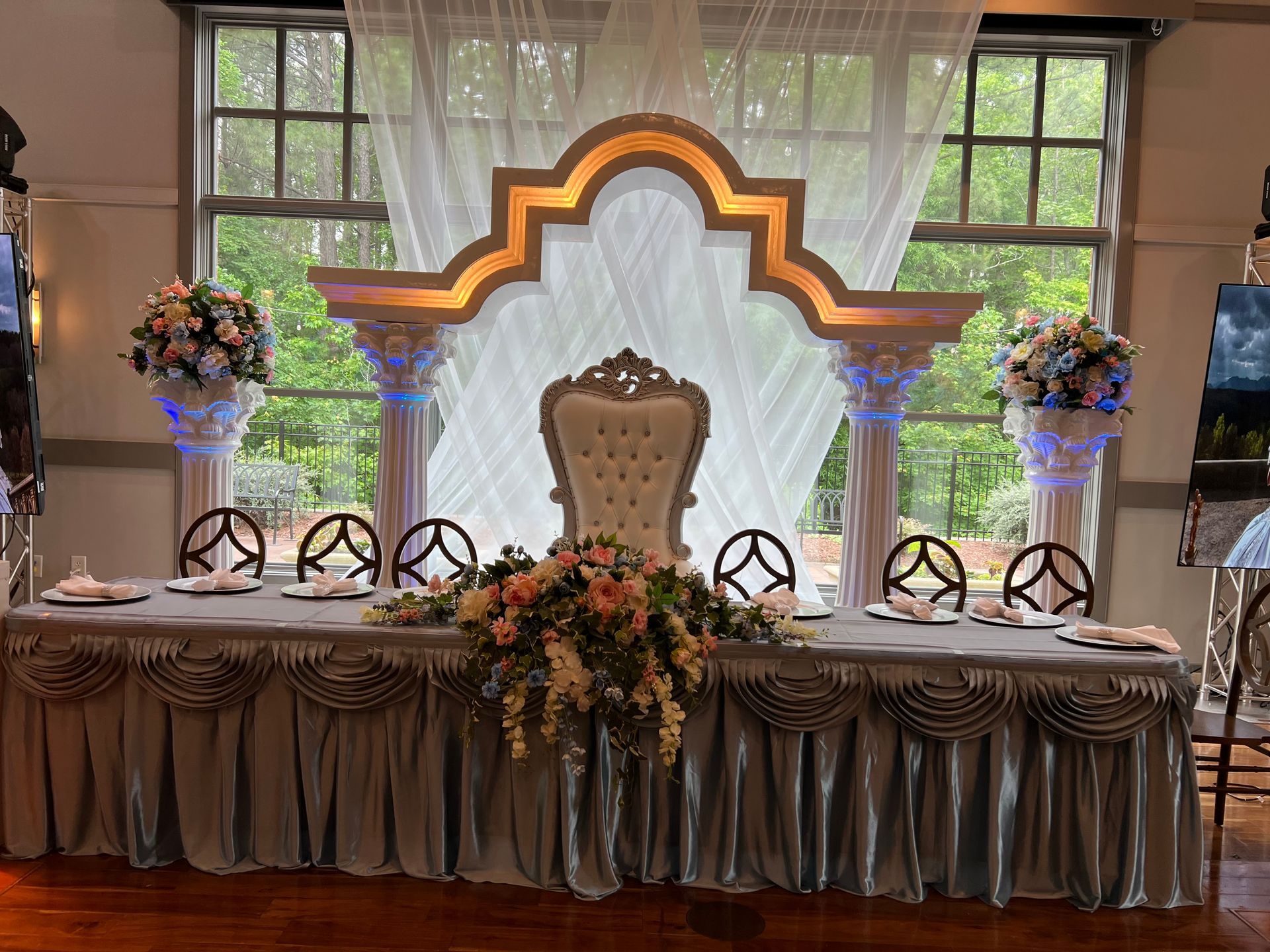 Decorated event table with ornate chair, floral arrangements, and glowing arched backdrop in front of windows