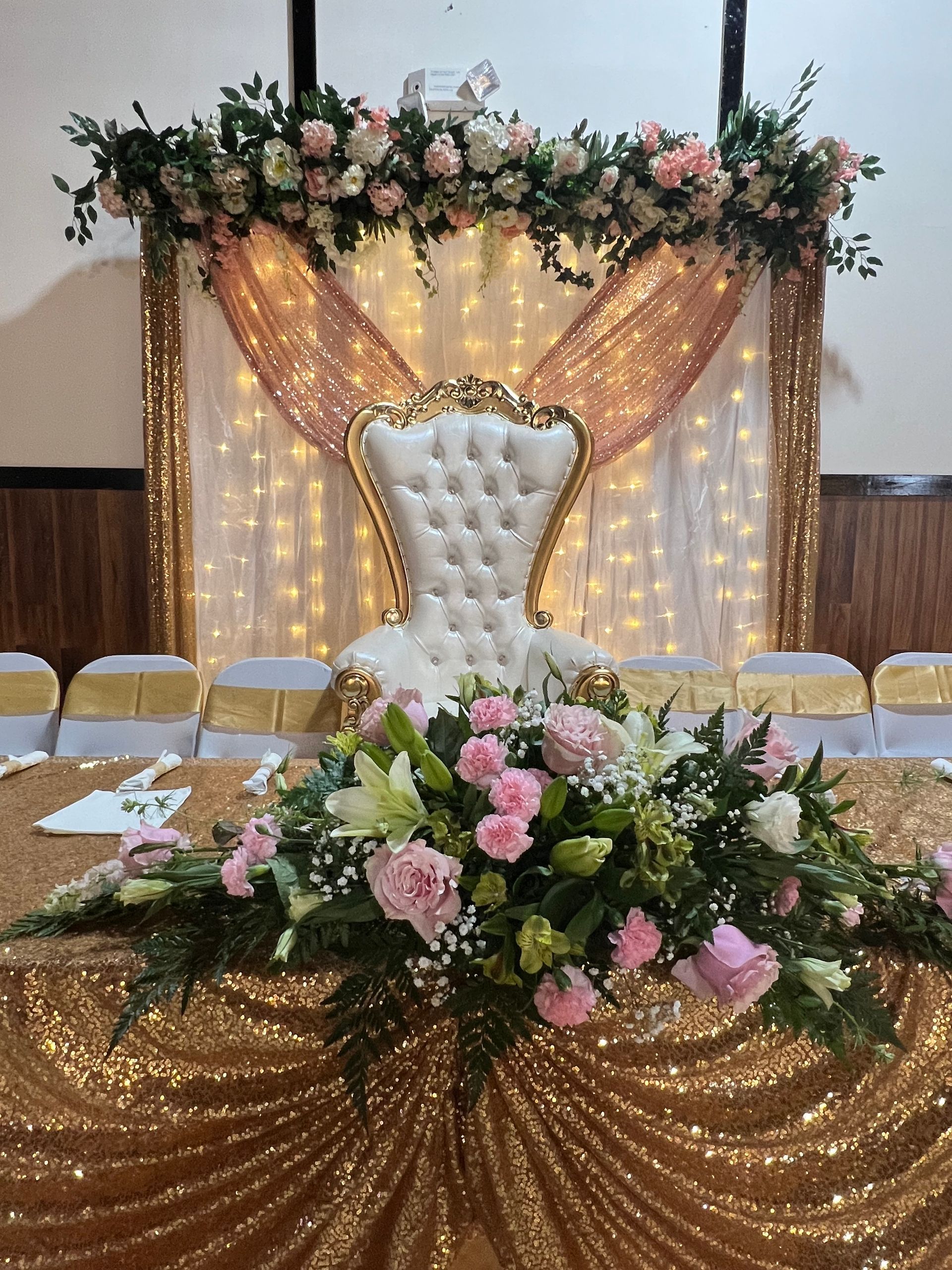 Wedding stage with white tufted chair, pink floral arrangements, and gold draped backdrop with lights