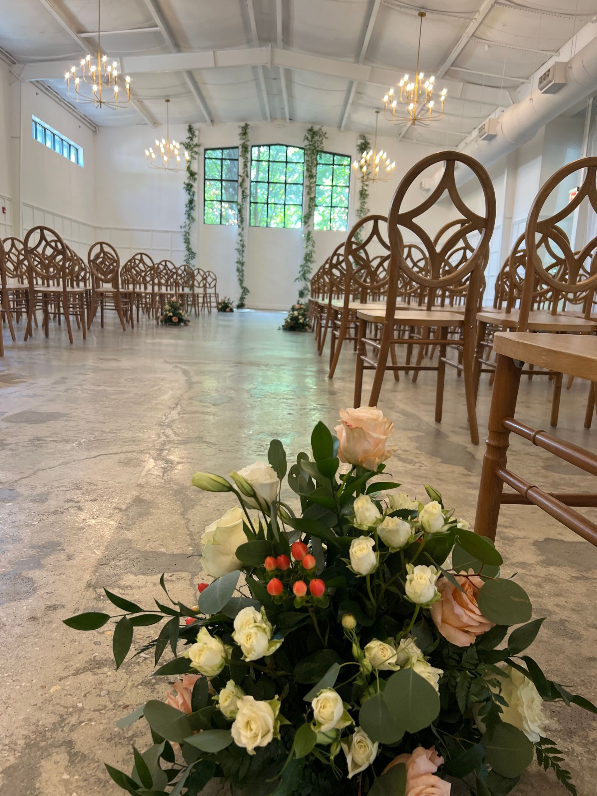 Floral arrangement in a bright ceremony hall with wicker chairs and chandeliers.