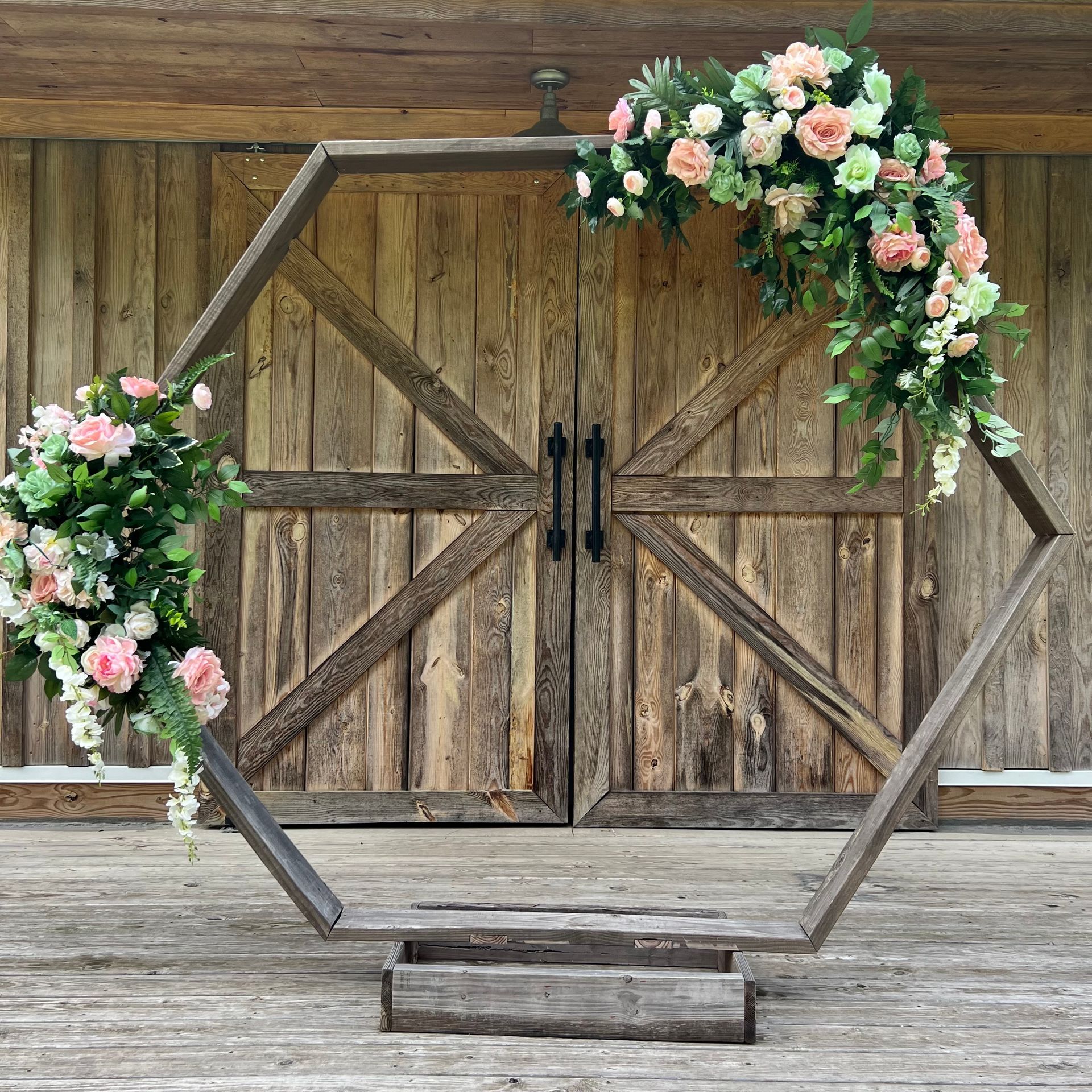 Hexagonal wooden wedding arch with pink and white flowers in front of rustic barn doors