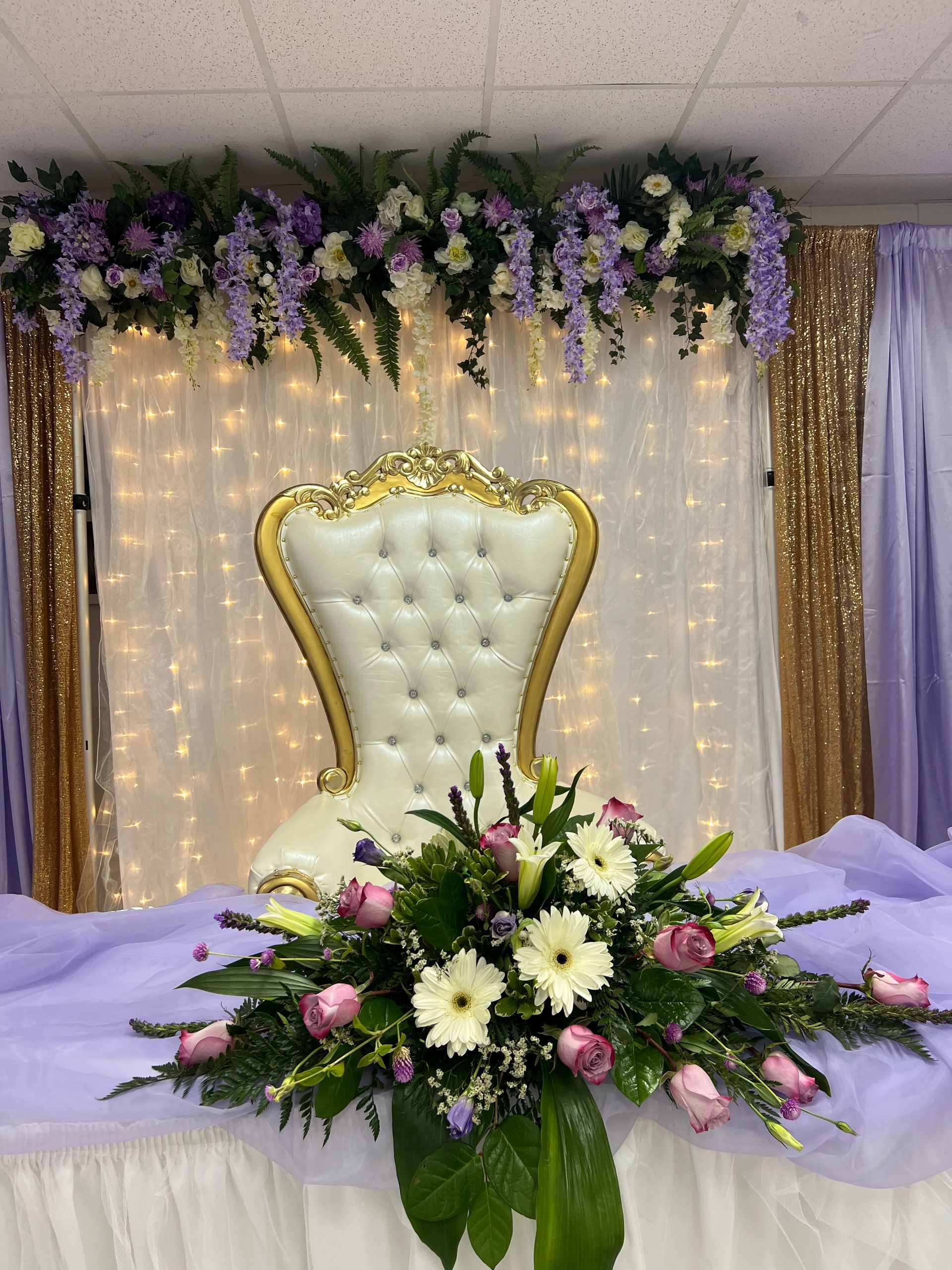Decorated wedding stage with gold-trimmed white chair, floral arrangement, and purple drapery with string lights