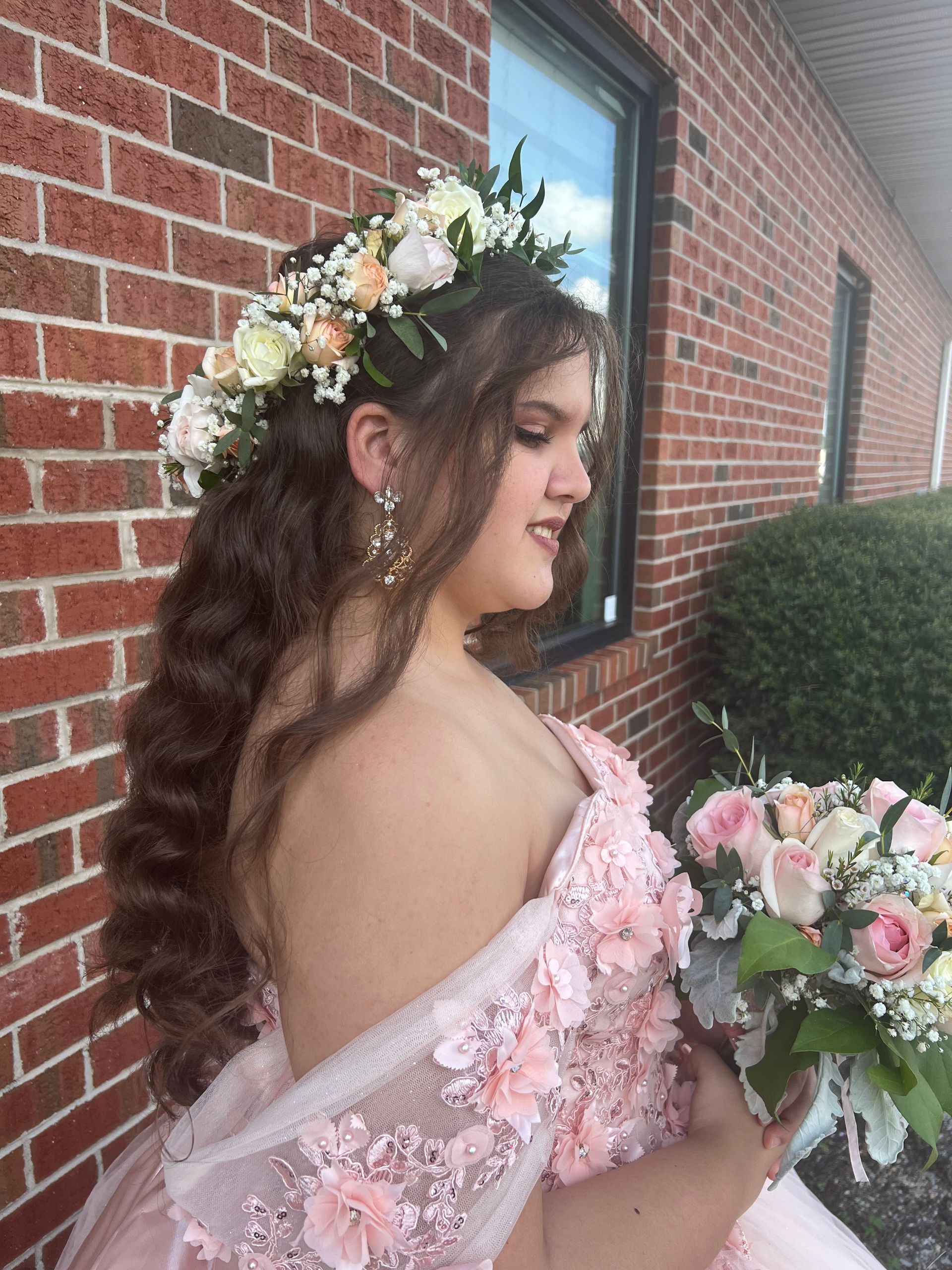 Young woman in a pink dress holding a bouquet, wearing a floral crown beside a brick wall