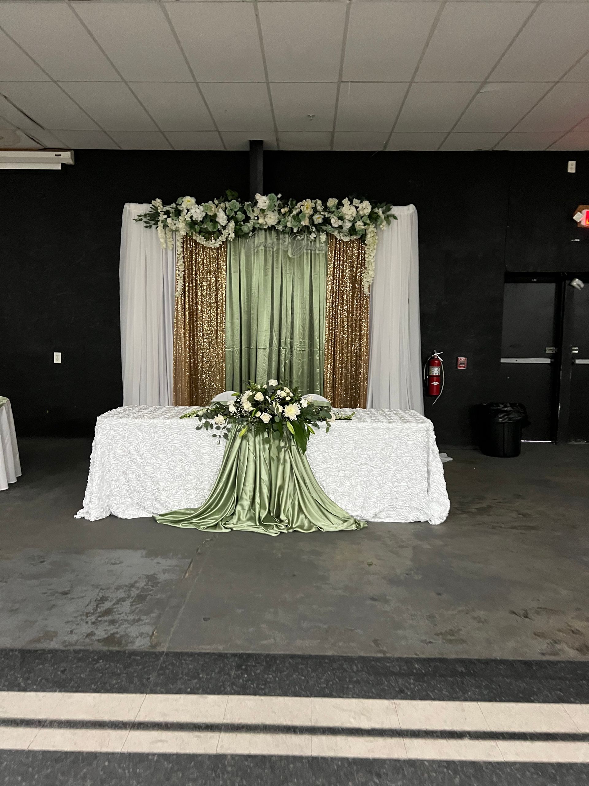 Decorated event backdrop with white, gold, and green drapes, floral garland, and white-covered tables