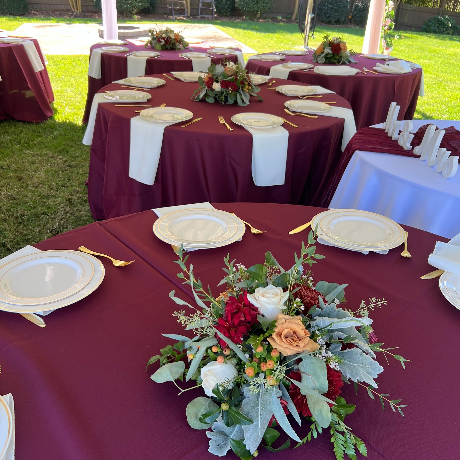 Round banquet tables with burgundy tablecloths and floral centerpieces set outdoors on a lawn.