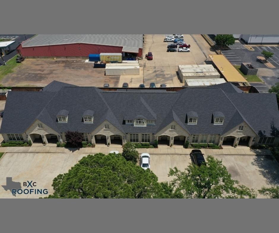 Aerial view of a large gray-roofed building with dormer windows and a parking lot behind it.