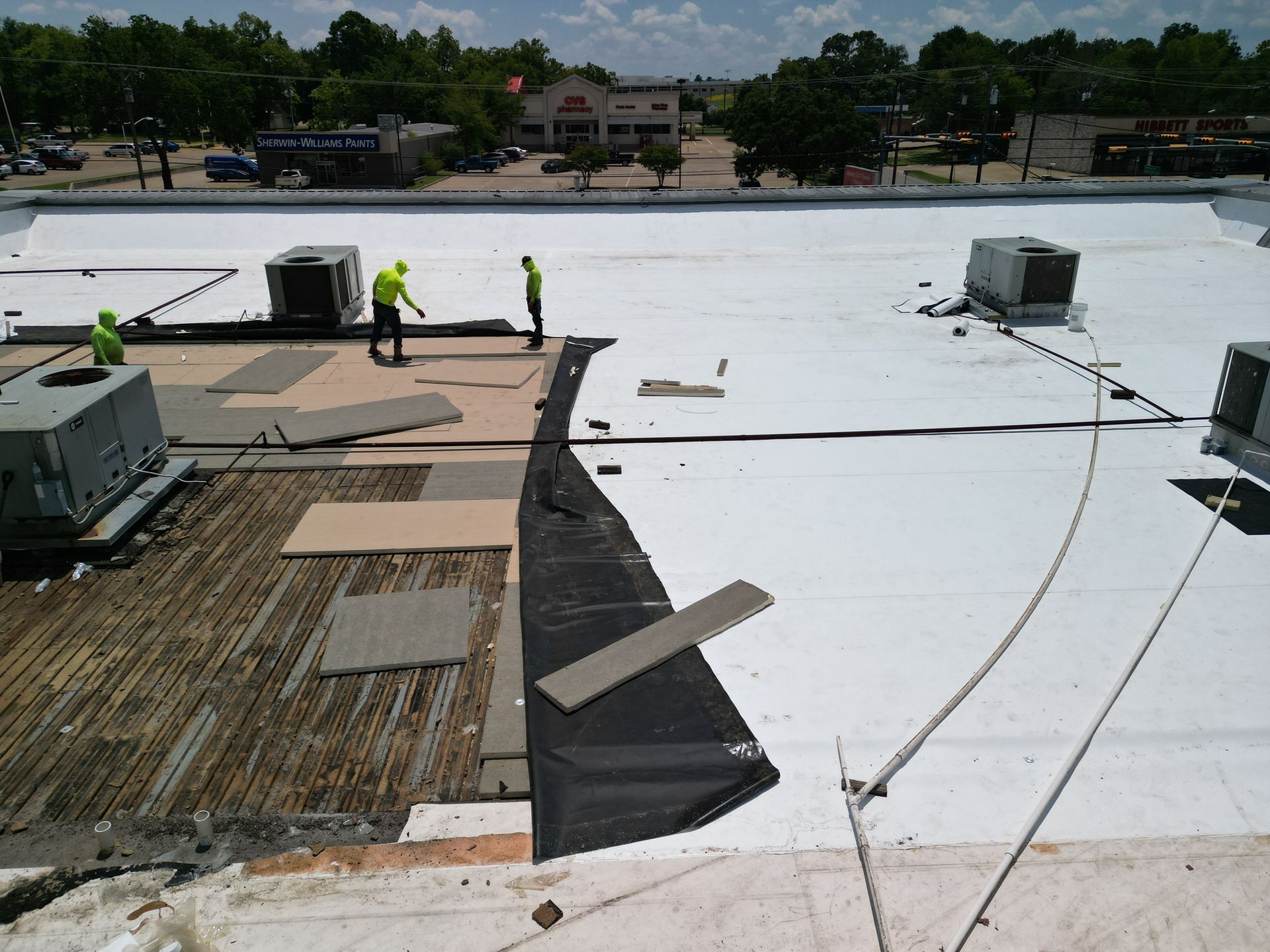 Workers on a flat roof installing black waterproofing beside white roofing, with equipment and rooftop vents.