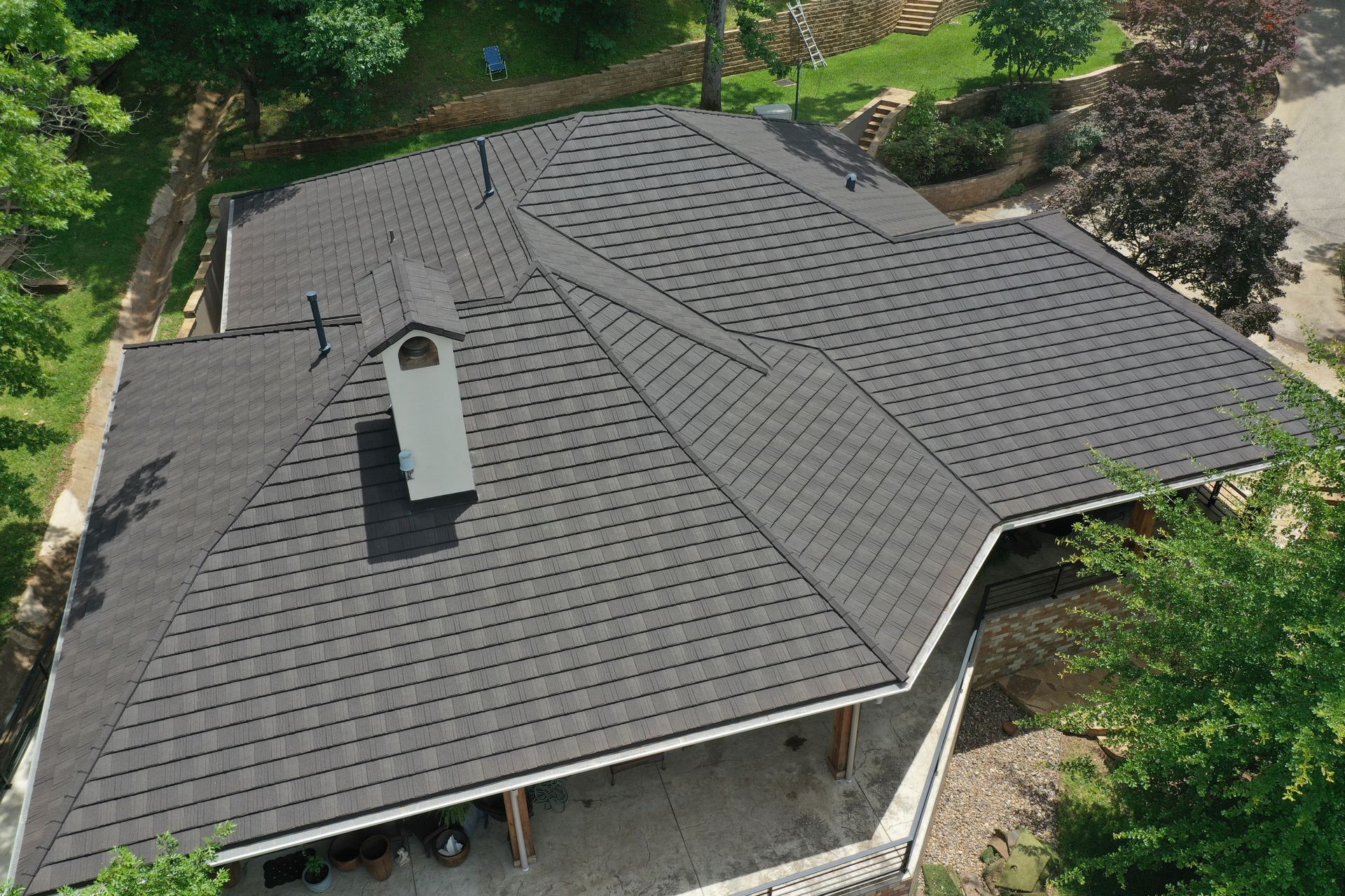 Aerial view of a gray shingled house roof with a chimney, surrounded by trees.