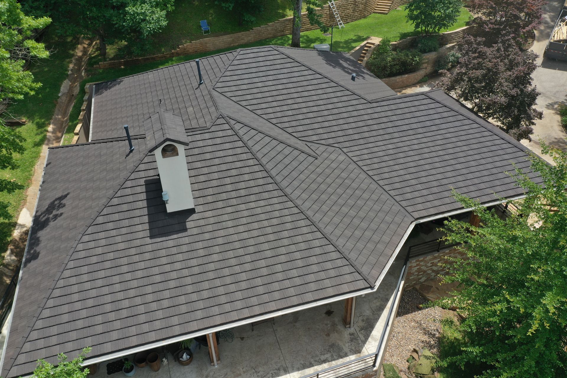 Aerial view of a gray shingled house roof with a chimney, surrounded by trees and greenery.