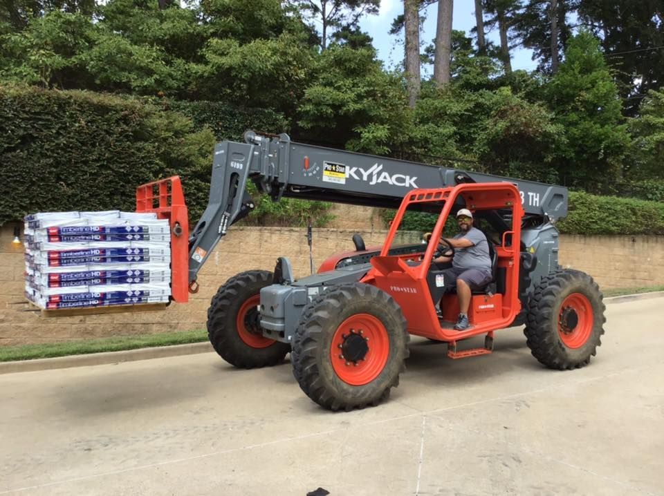 Orange forklift carrying stacked pallets on a paved road near trees and a building