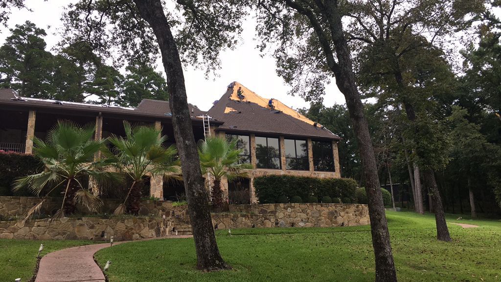 Large house with stone foundation and dark roof, surrounded by tall trees and a green lawn.