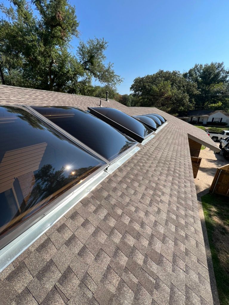 Curved rooftop skylights on a shingled roof under a clear blue sky, with trees and a patio below