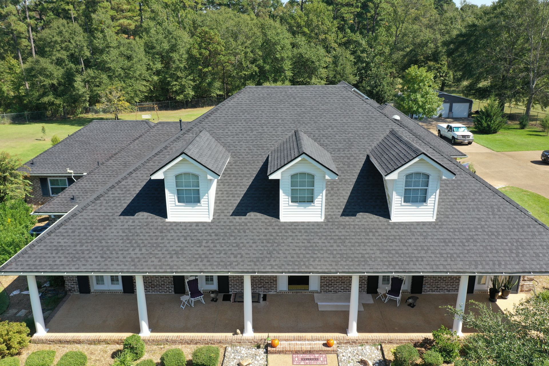 Aerial view of a gray-roofed house with dormer windows and a covered porch surrounded by trees.